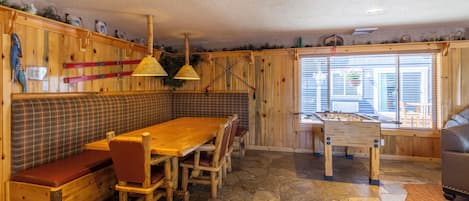 A cozy wooden interior with a stone floor, a wooden table with chairs, a bench, and a foosball table.