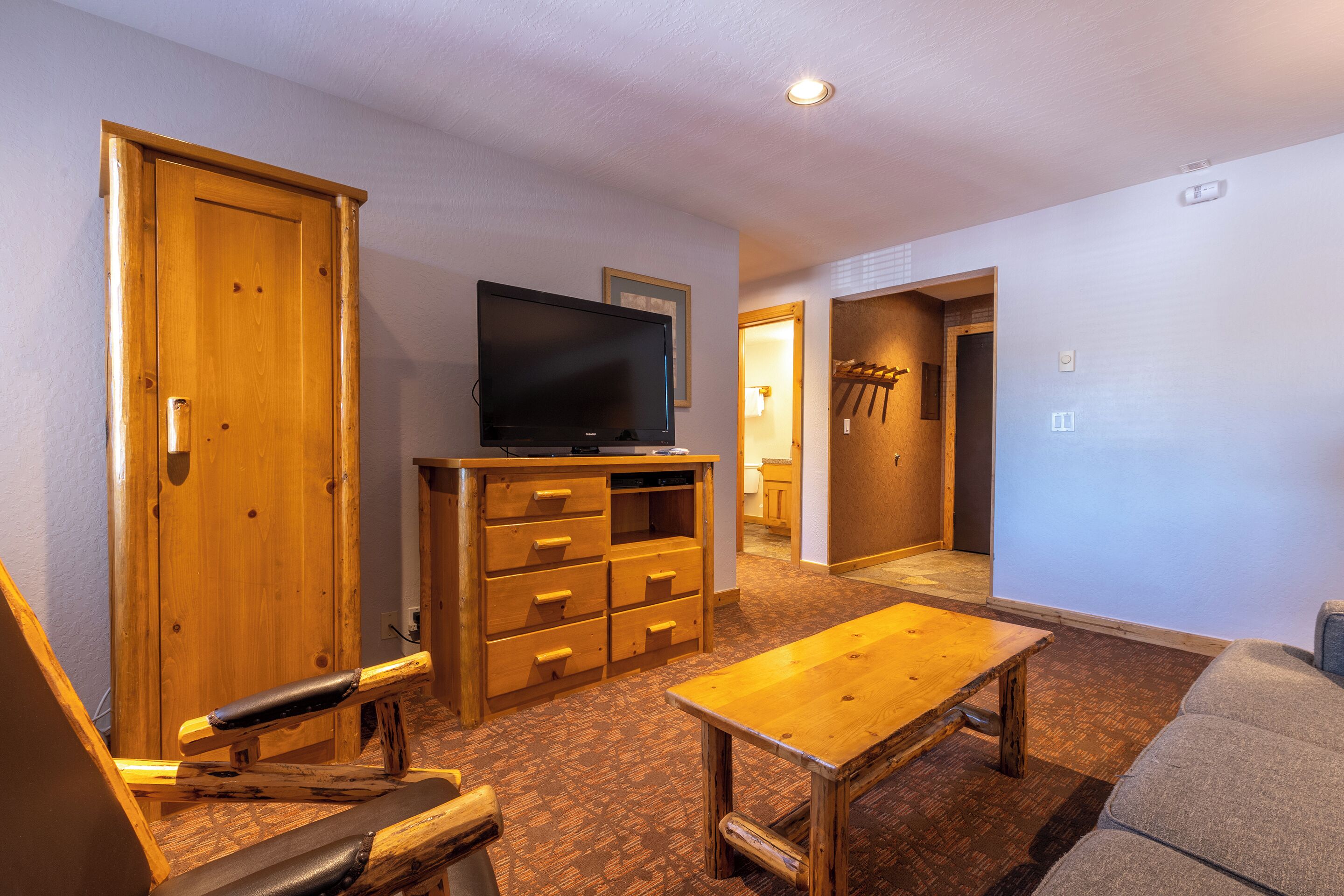 A room with a wooden bench, a television on a wooden stand, a wooden table, and a door leading to another room.