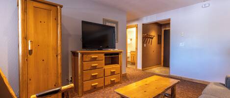 A room with a wooden bench, a television on a wooden stand, a wooden table, and a door leading to another room.