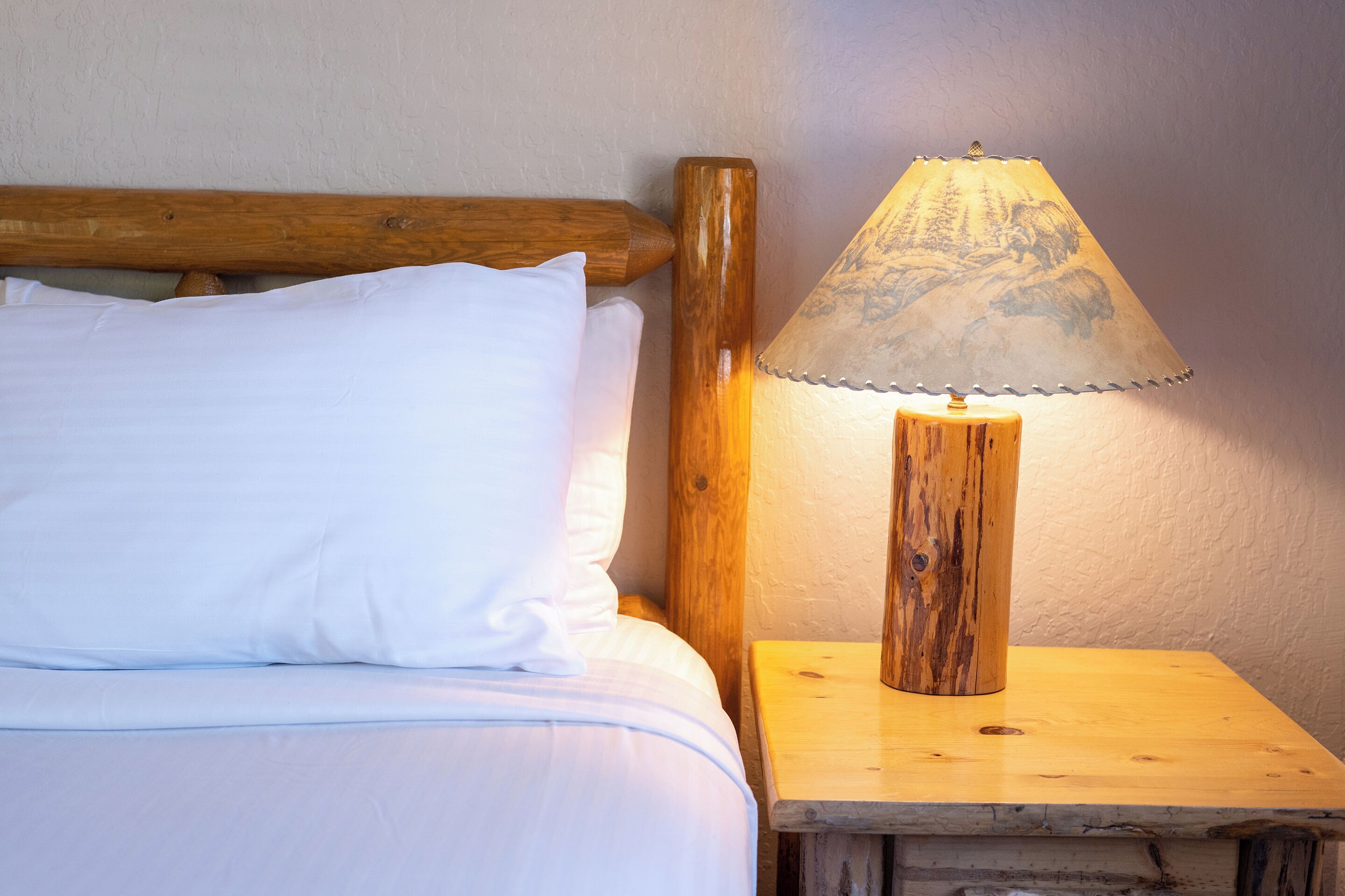 A wooden bedside table with a lamp, a bed with white bedding, and a wooden headboard.