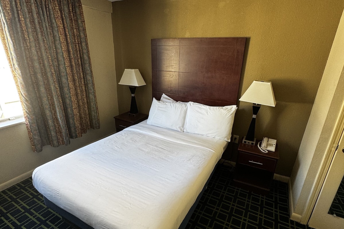 A hotel room with a large bed, two bedside tables with lamps, a patterned carpet, and a window with curtains.