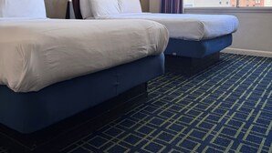 Habitación de hotel con dos camas, un ventanal amplio con vista a la ciudad, una alfombra estampada y una lámpara.