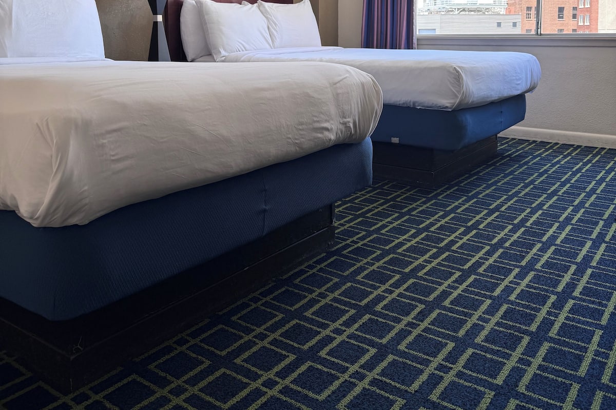 A hotel room with two beds, a large window with a city view, a patterned carpet, and a lamp.