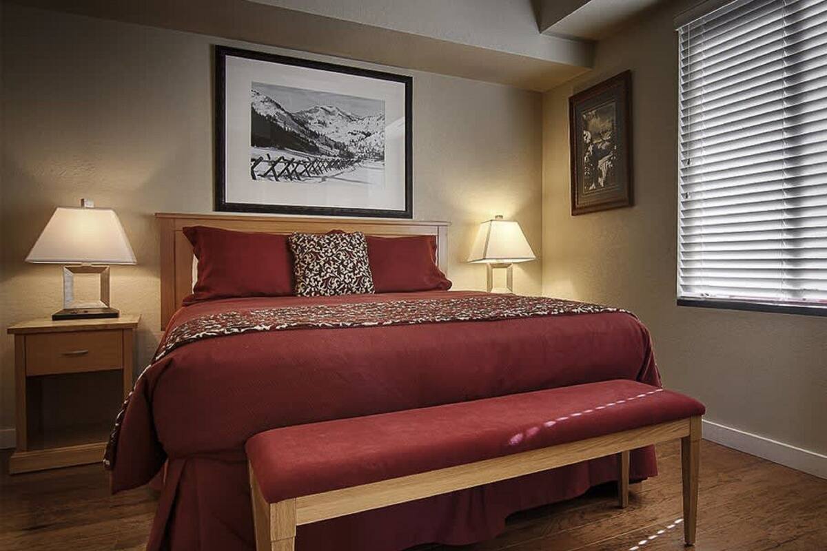 Quarto com uma cama grande, duas mesinhas de cabeceira, um banco e duas luminárias fixadas na parede.