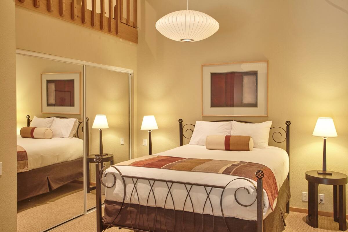 Quarto de hotel com uma cama grande, duas mesas de cabeceira com luminárias, um espelho, um quadro na parede e uma escada de madeira.