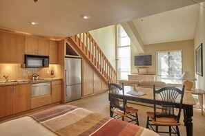A kitchen with wooden cabinets, a microwave, and a refrigerator. There is a dining area with a table and chairs, and a staircase leading to another level.