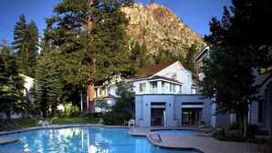 A swimming pool with a mountain backdrop, surrounded by trees and a building with a white facade.