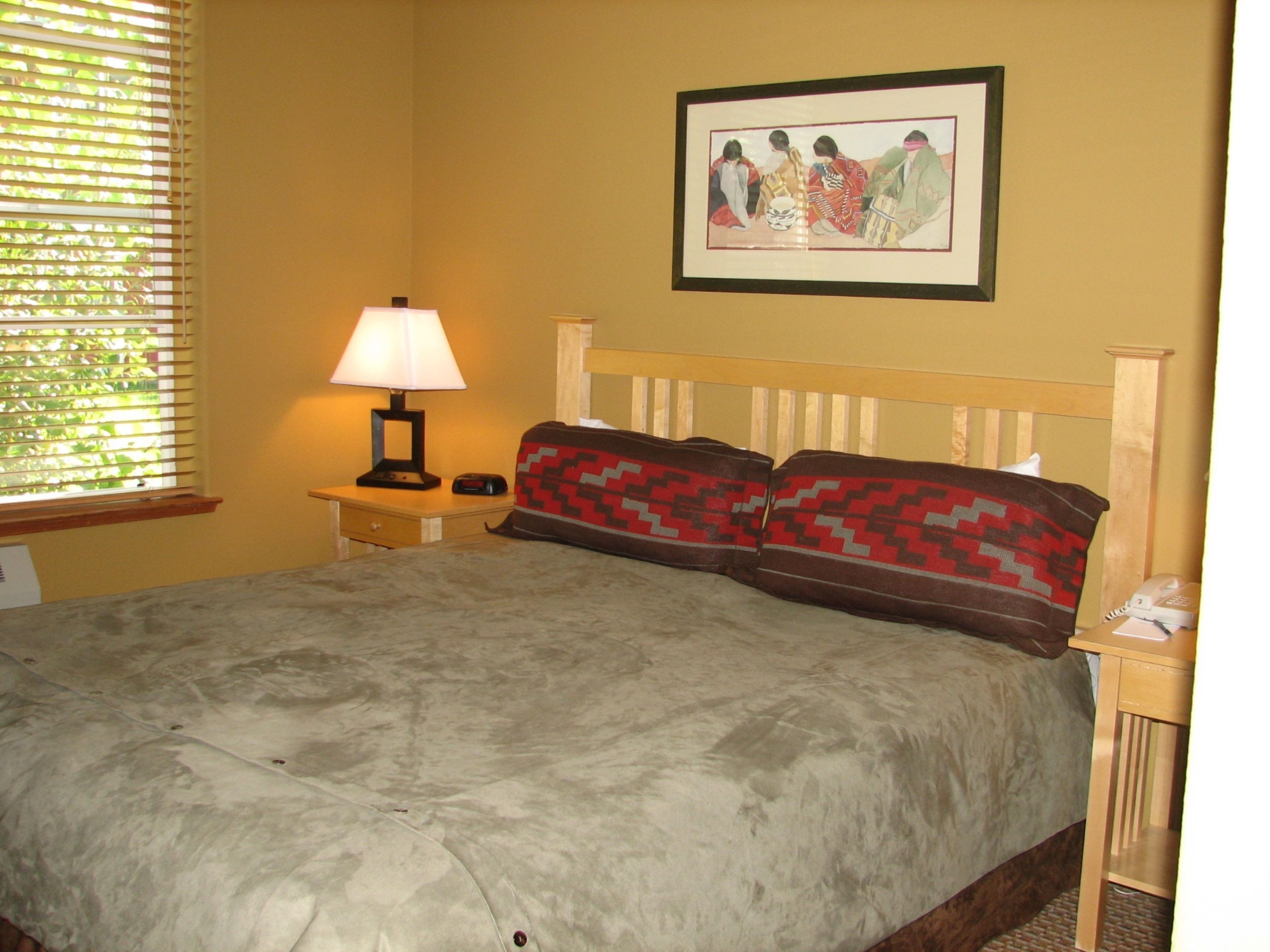 A bedroom with a bed, a nightstand, a lamp, a framed picture on the wall, and a window with blinds.