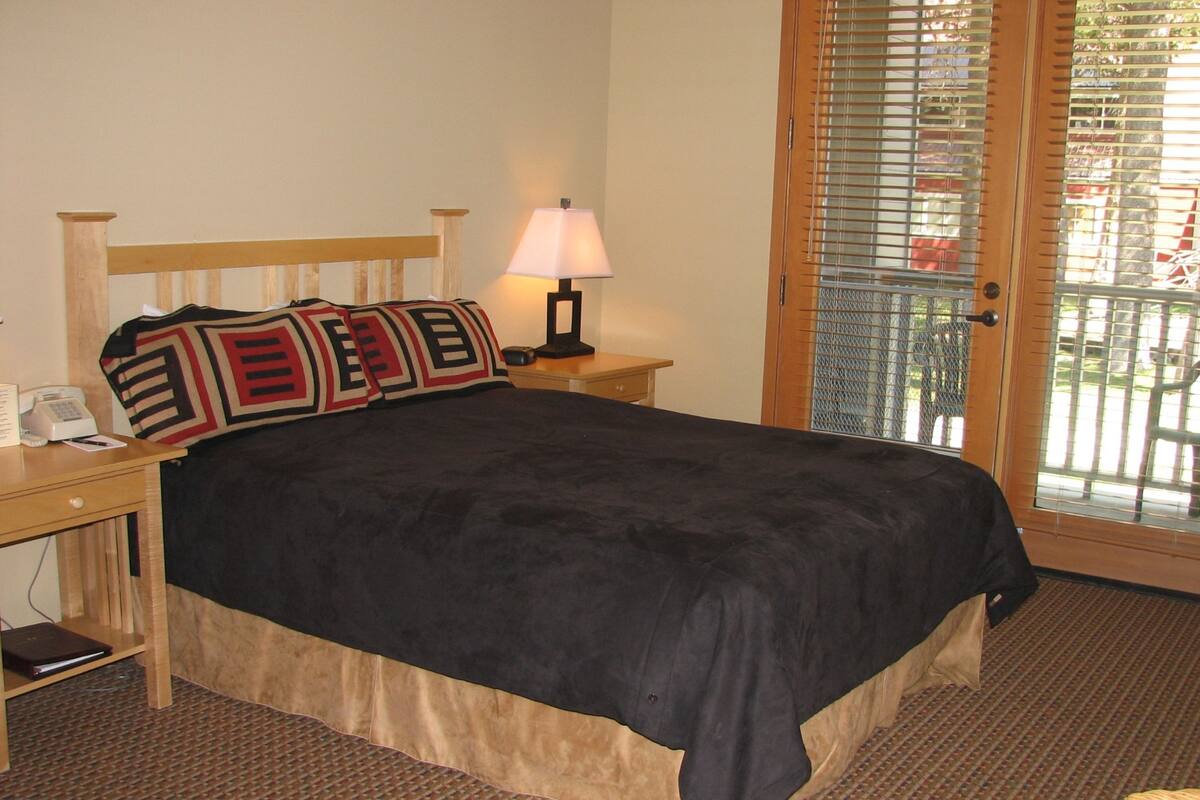 Quarto de hotel com cama, mesa de cabeceira, abajur e vista pela porta.