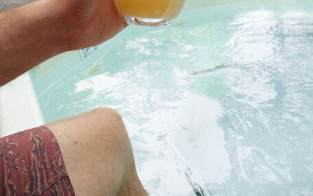 A person holding a glass of beer near a pool with lounge chairs and umbrellas in the background.