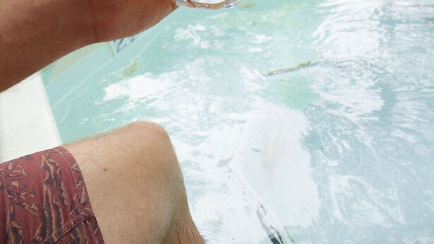 Una persona sostiene un vaso de cerveza cerca de una piscina con sillones de descanso y sombrillas al fondo.