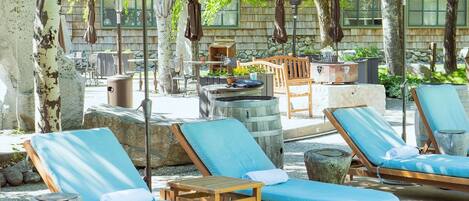 Zona junto a la piscina con sillas de descanso y sombrillas, rodeada de árboles y construcciones de piedra.