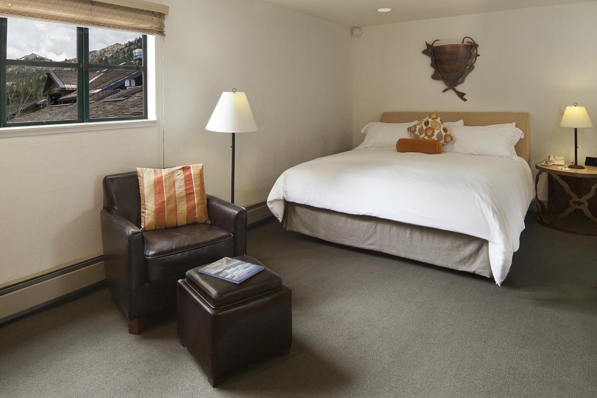 A hotel room with a large bed, a leather chair, a small ottoman, and a view of buildings through a window.