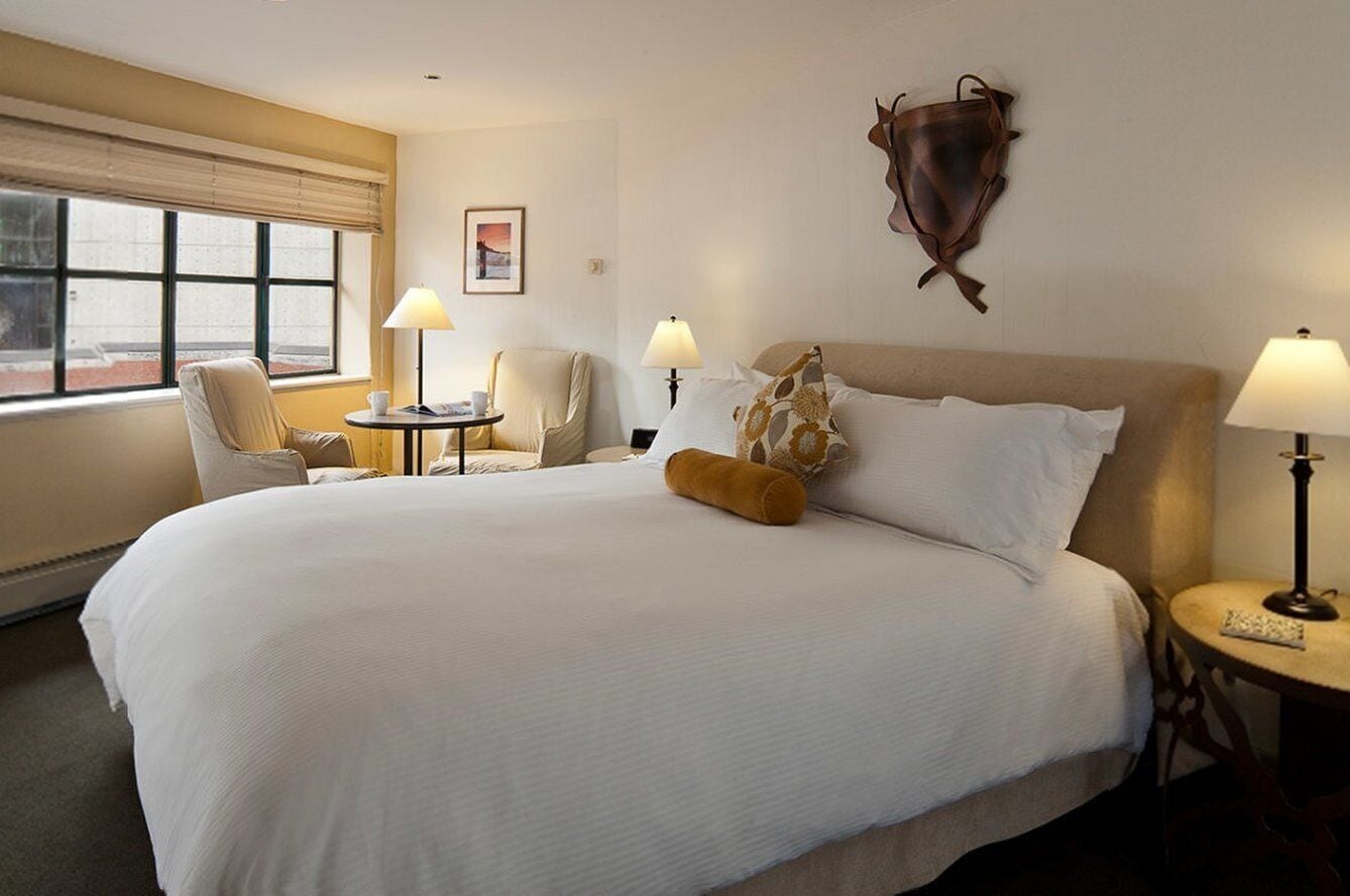 Une chambre d’hôtel avec un grand lit, deux tables de chevet avec des lampes, une chaise et une fenêtre avec des stores.