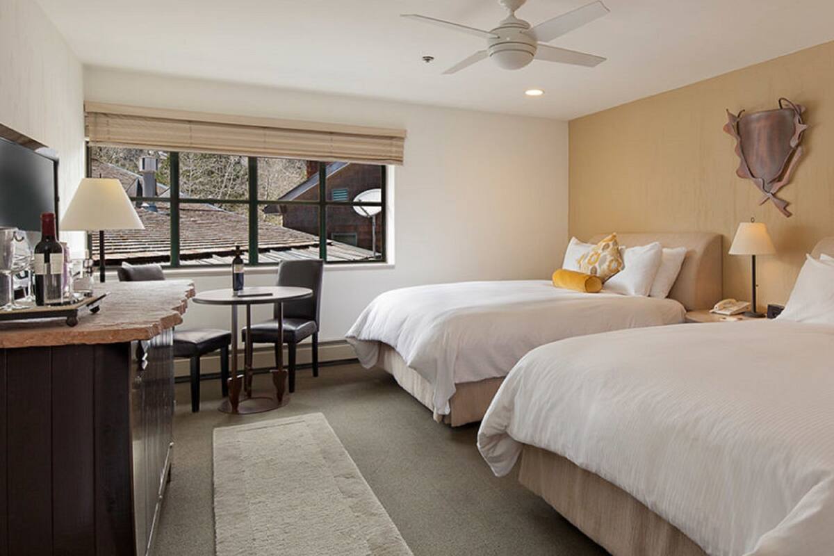 A hotel room with two beds, a desk with a chair, a small table, and a window with a view of the outside.