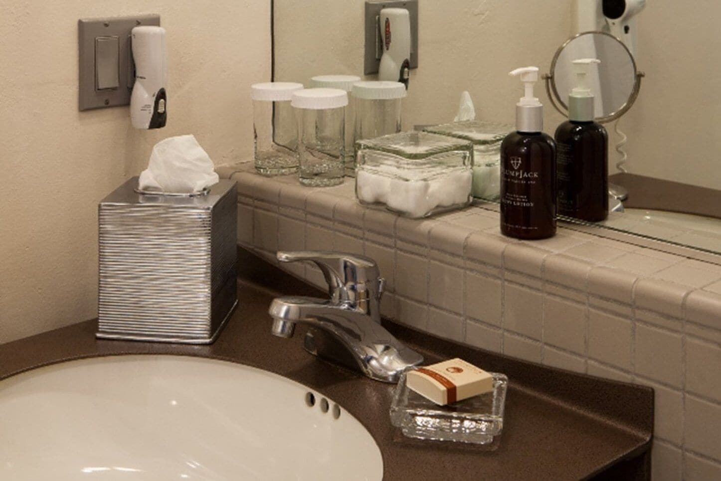 A bathroom with a sink, mirror, toiletries, and a tissue dispenser.