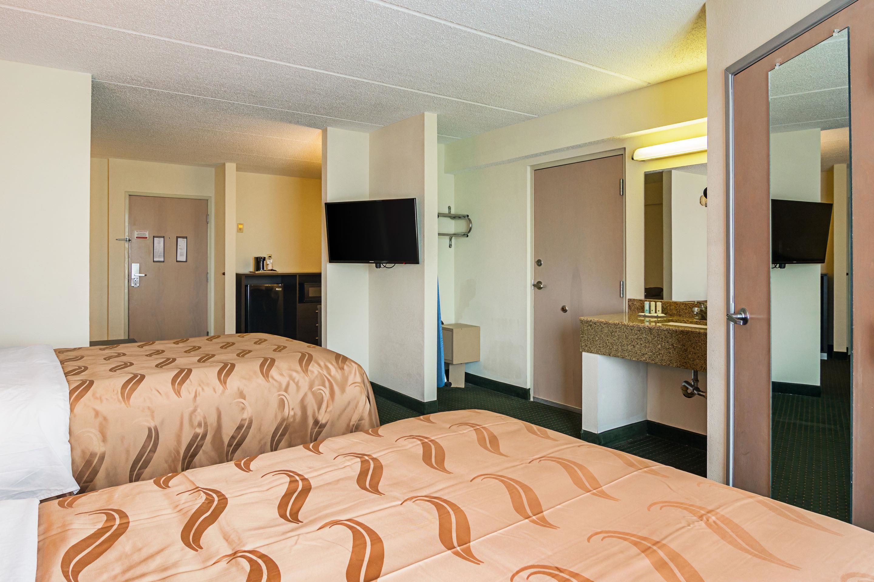 A hotel room with two beds, a television, a desk, and a bathroom with a sink and mirror.
