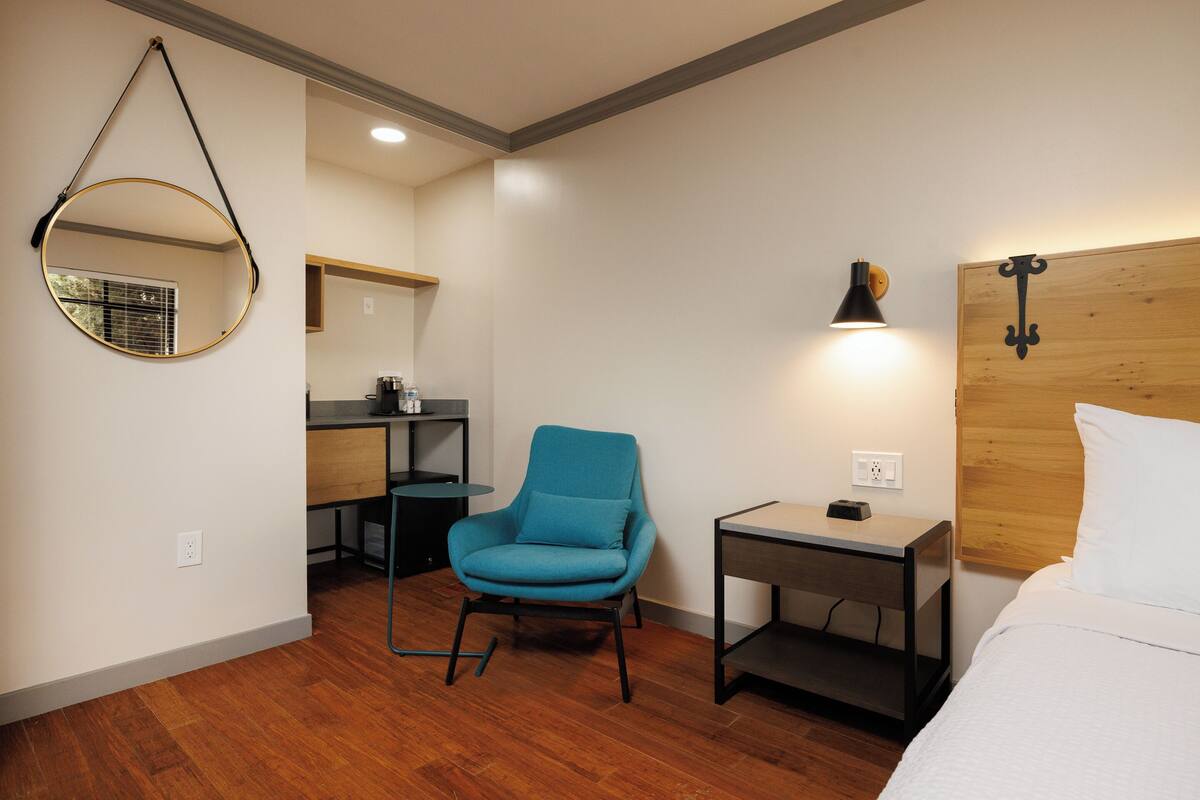 A hotel room with a wooden floor, a blue armchair, a bedside table with a lamp, a bed with white bedding, and a round mirror on the wall.