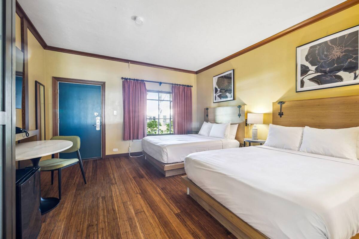 A hotel room with two beds, a dining table, a chair, a window with curtains, a wall-mounted lamp, and a framed picture.