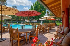 A poolside area with wooden tables and chairs, umbrellas, and a pool.