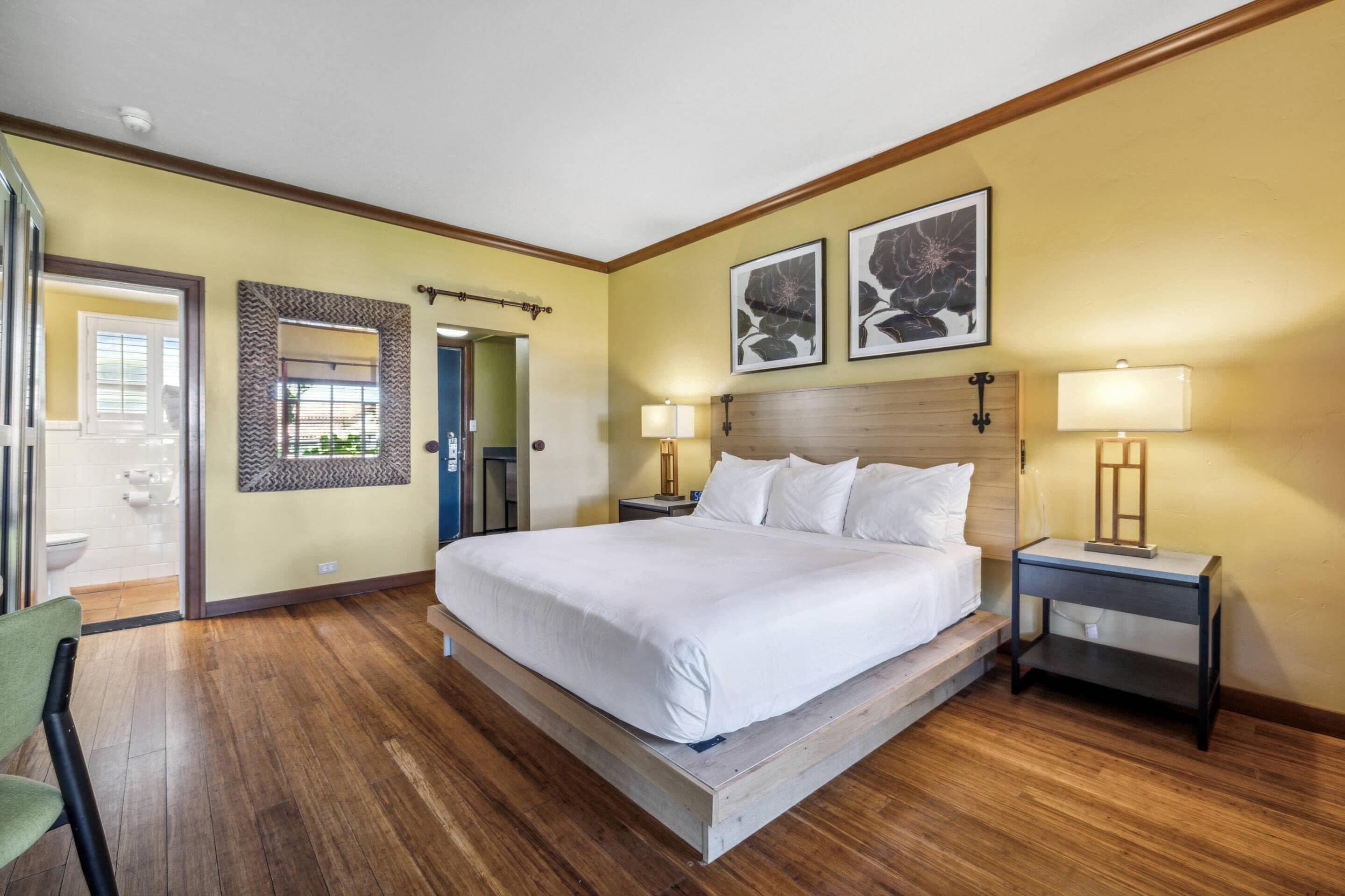 Habitación de hotel con una cama grande, dos mesitas de noche con lámparas, un cabecero de madera, un espejo y una puerta que da a un baño.