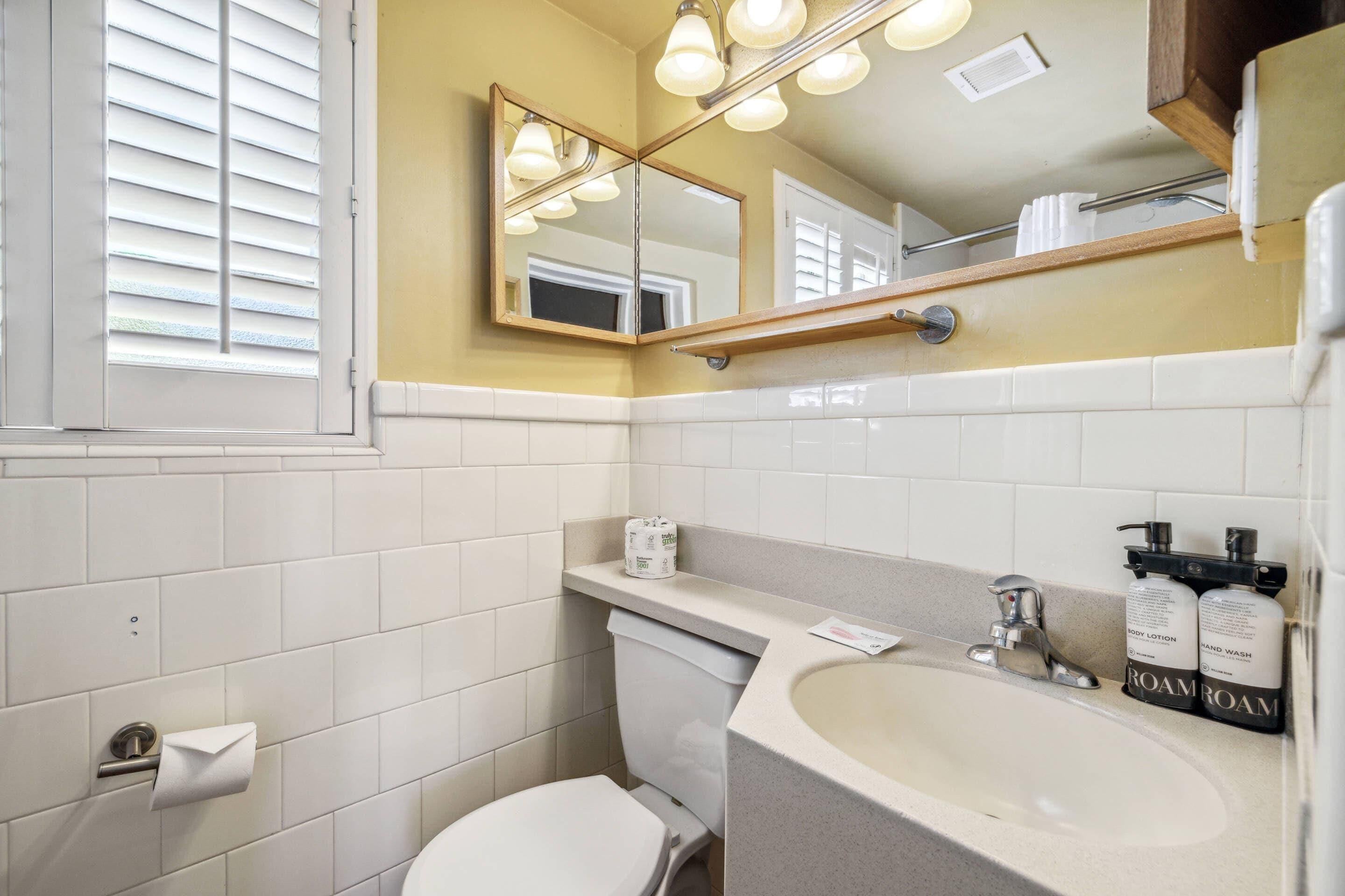 Un baño con azulejos blancos, un inodoro, un lavabo con un espejo encima y dos botellas de loción corporal.