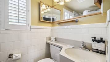 Un baño con azulejos blancos, un inodoro, un lavabo con un espejo encima y dos botellas de loción corporal.