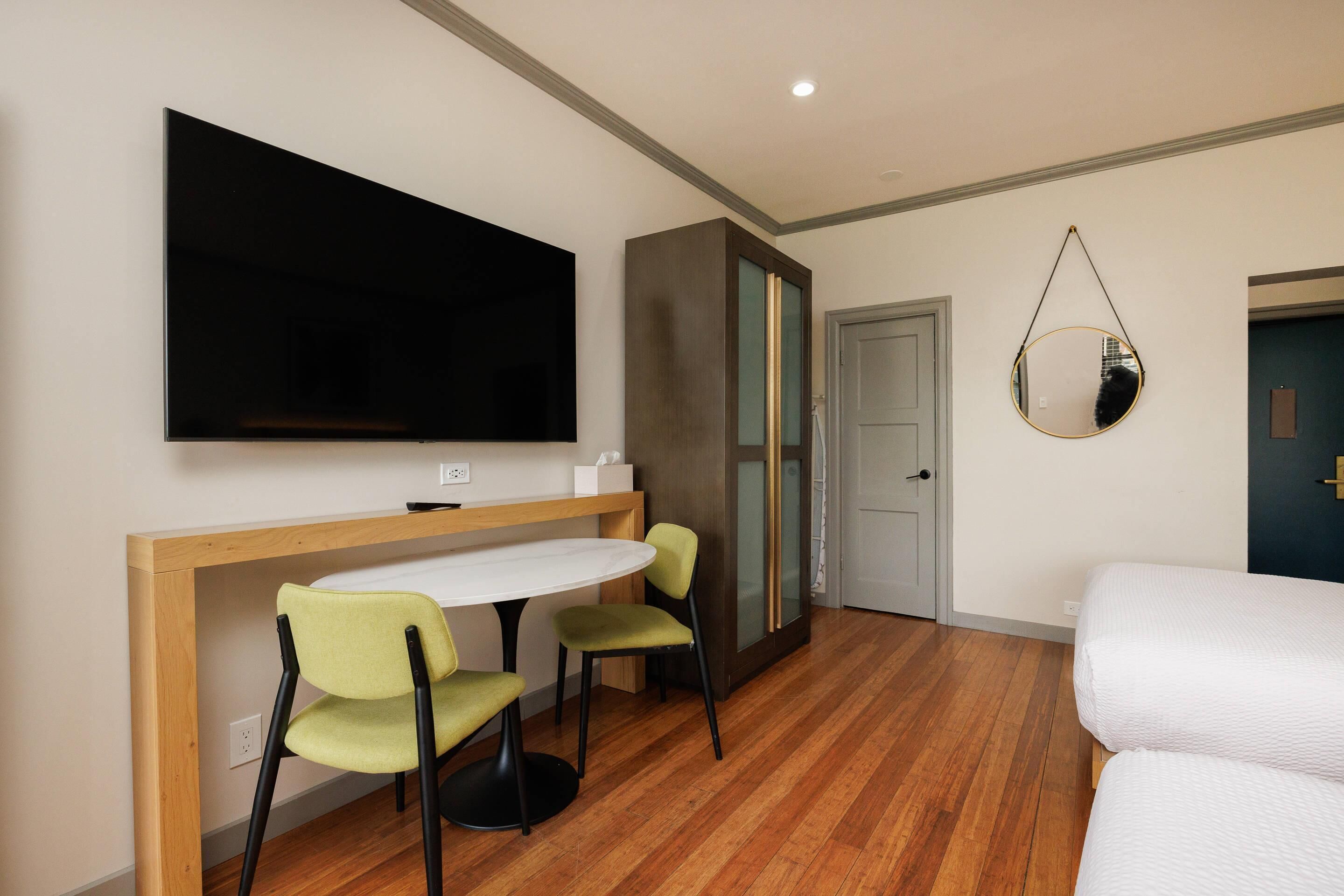 Quarto com televisão, secretária com cadeiras, cama e espelho na parede.