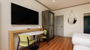 Quarto com televisão, secretária com cadeiras, cama e espelho na parede.