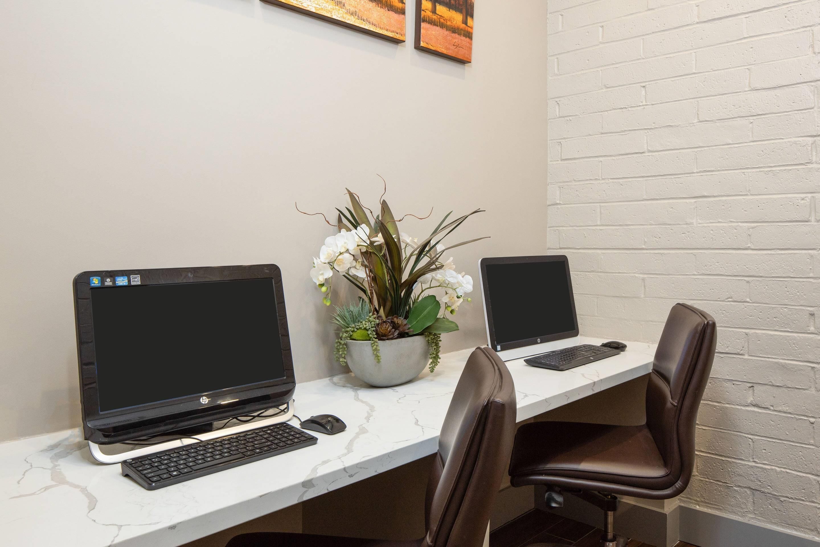 Two laptops on a white marble desk with a potted plant, two black chairs, and a white brick wall.