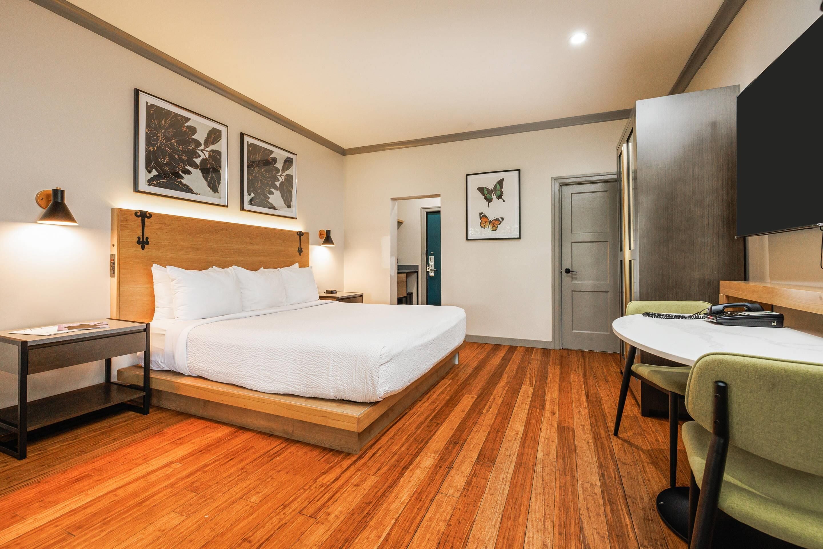 Ein Hotelzimmer mit Holzboden, einem Bett mit weißer Bettwäsche, einem Schreibtisch mit Stuhl und Bildern an den Wänden.