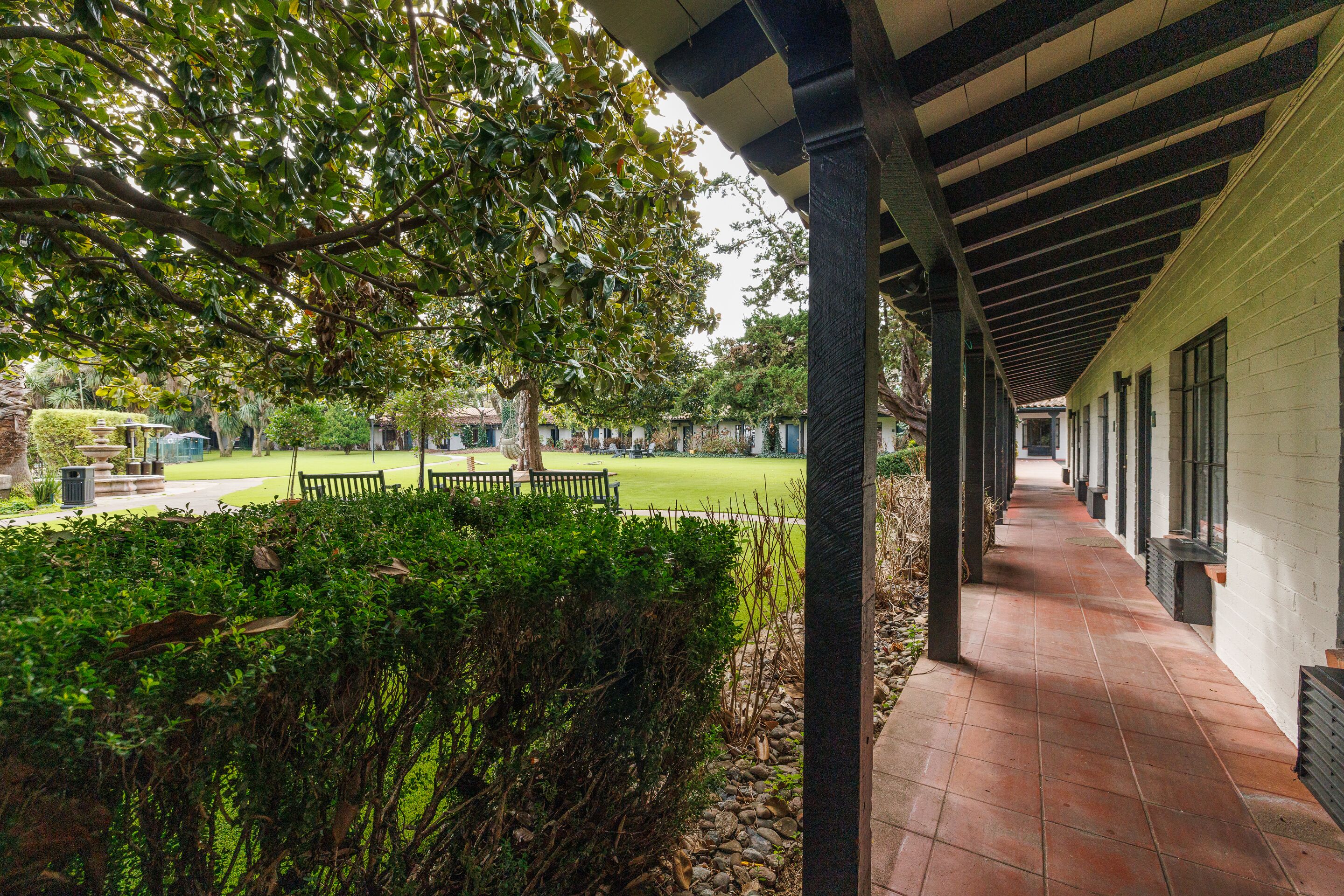 A covered walkway with a tiled floor, leading to a grassy area with benches and a tree.