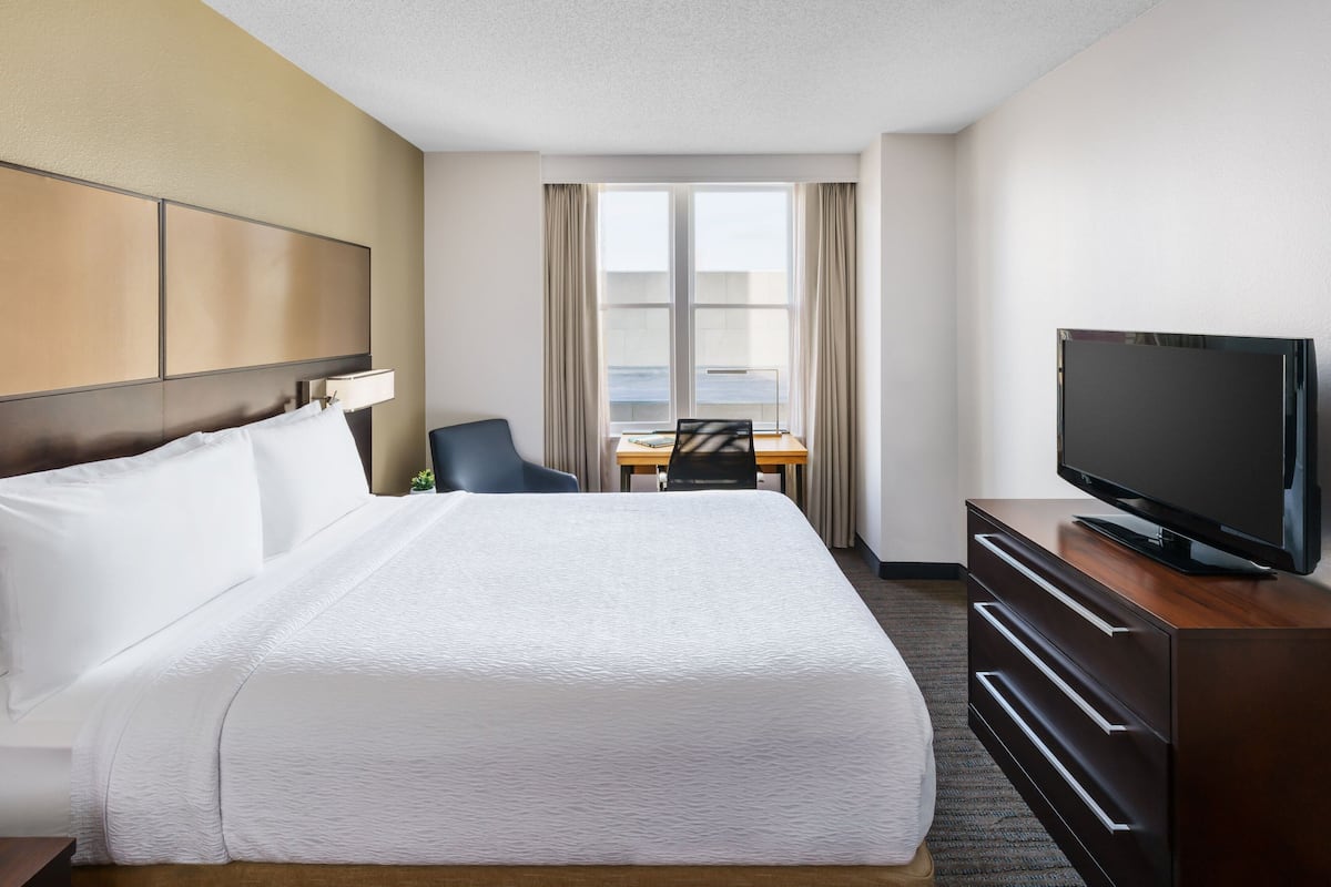 A hotel room with a large bed, a TV on a wooden dresser, a desk with a chair, and a window with curtains.