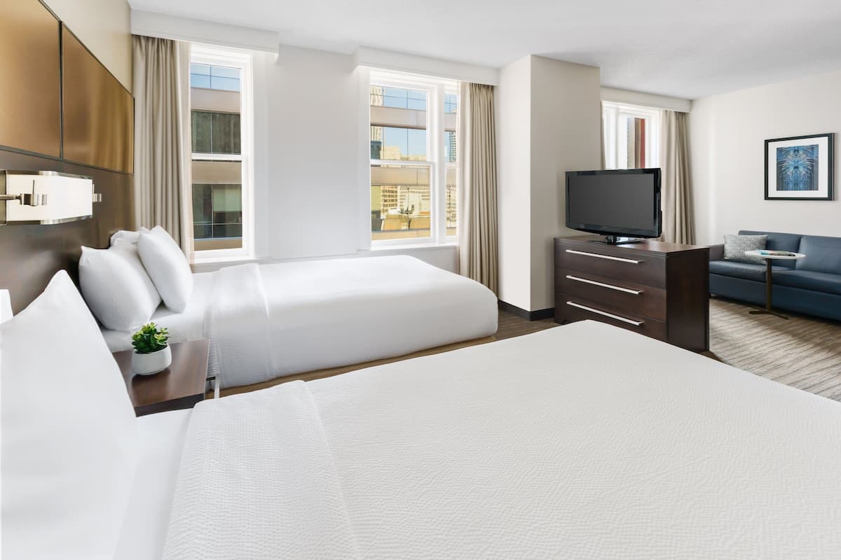 A hotel room with two beds, a TV, a sofa, and a window with a view of buildings.