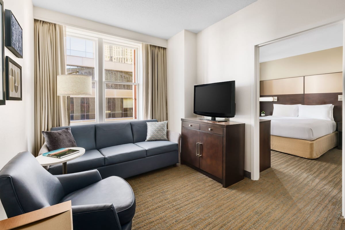 A modern hotel room with a sofa, a television, a bed, and a view of the city through large windows.