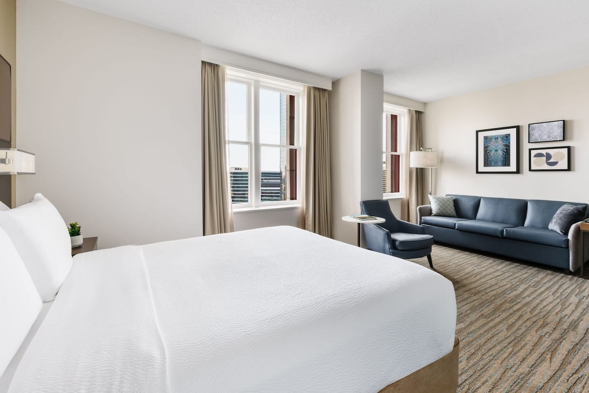 A hotel room with a large bed, a sofa, a small table, and a chair. There are curtains on the windows, and a lamp on the table.