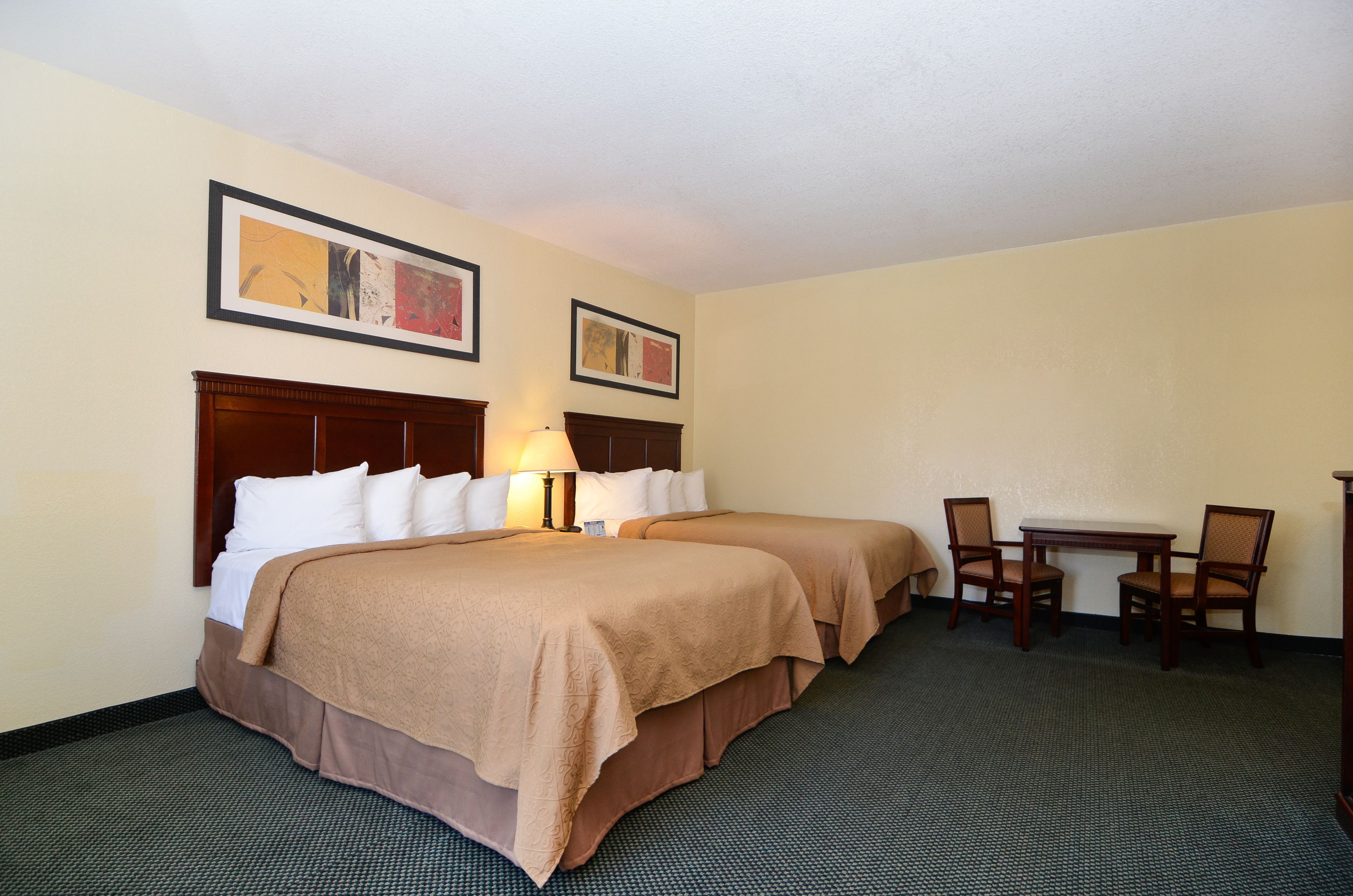 A hotel room with two beds, a desk, and chairs. There are framed pictures on the wall.