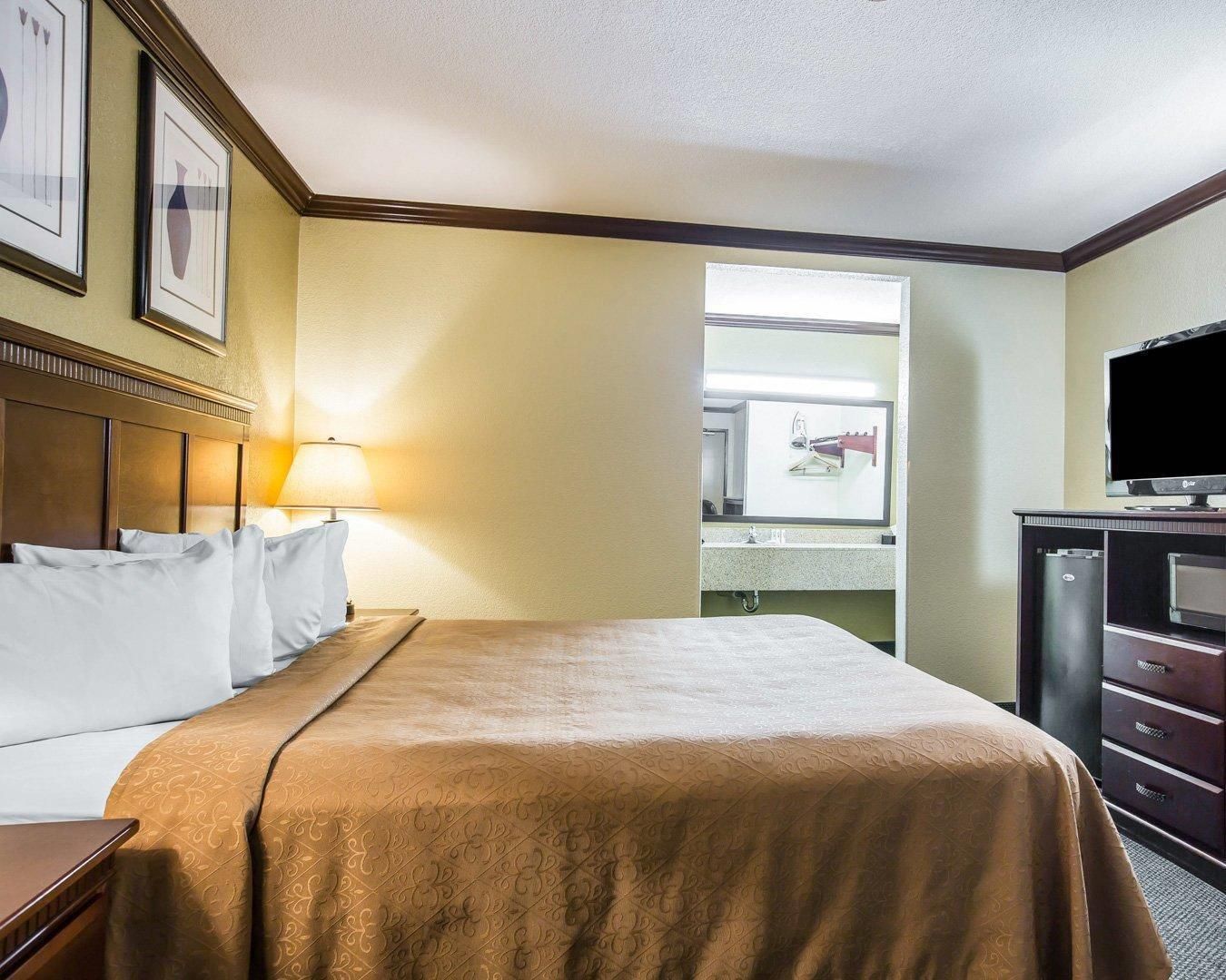 Une chambre d’hôtel avec un lit, un bureau, une lampe, une télévision et une salle de bain visible à travers une porte ouverte.
