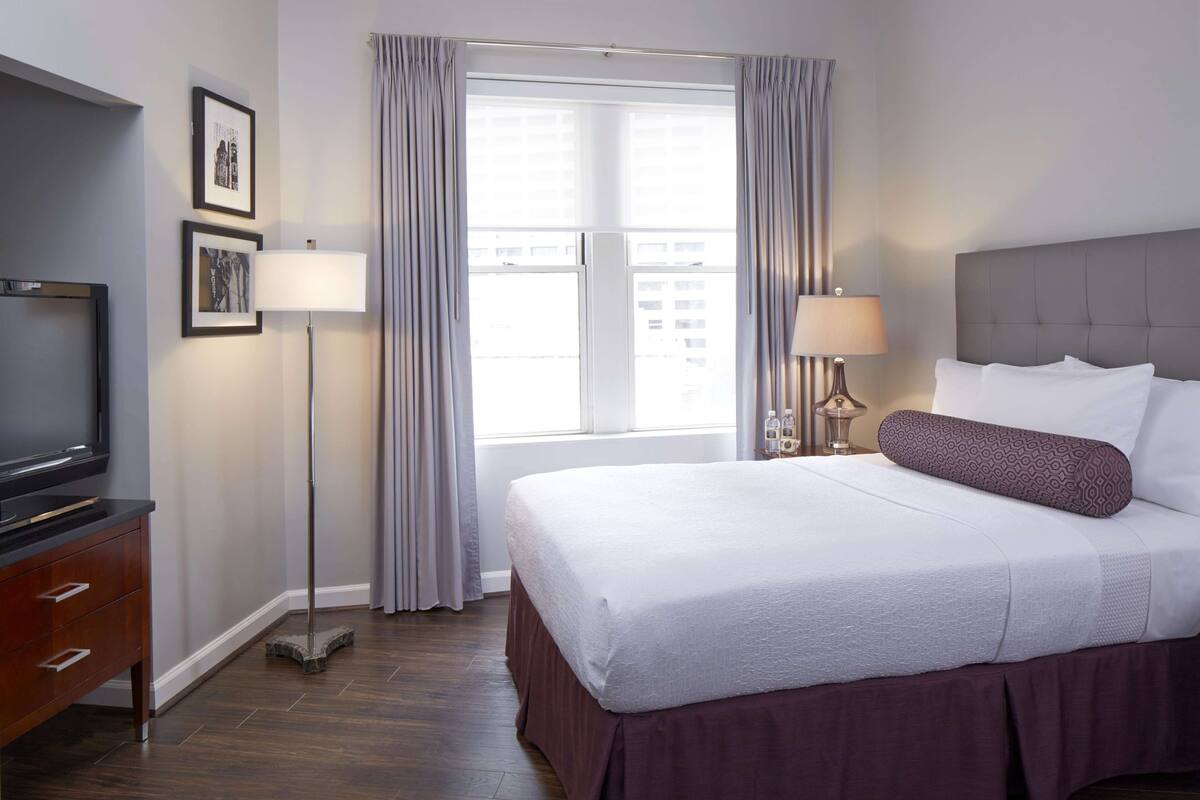 A hotel room with a bed, a television, a floor lamp, and a window with curtains.