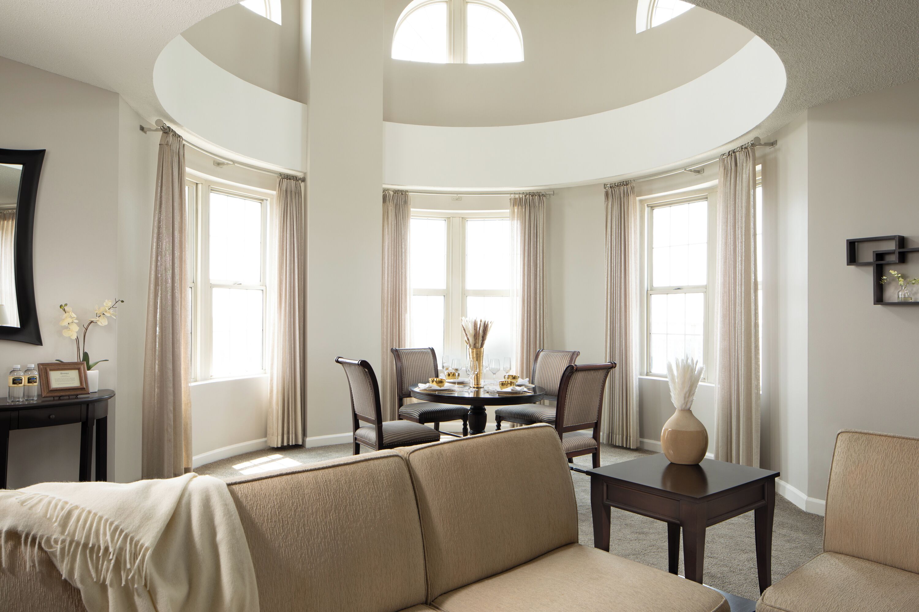 A spacious living room with a round dining table, comfortable seating, and large windows with curtains.