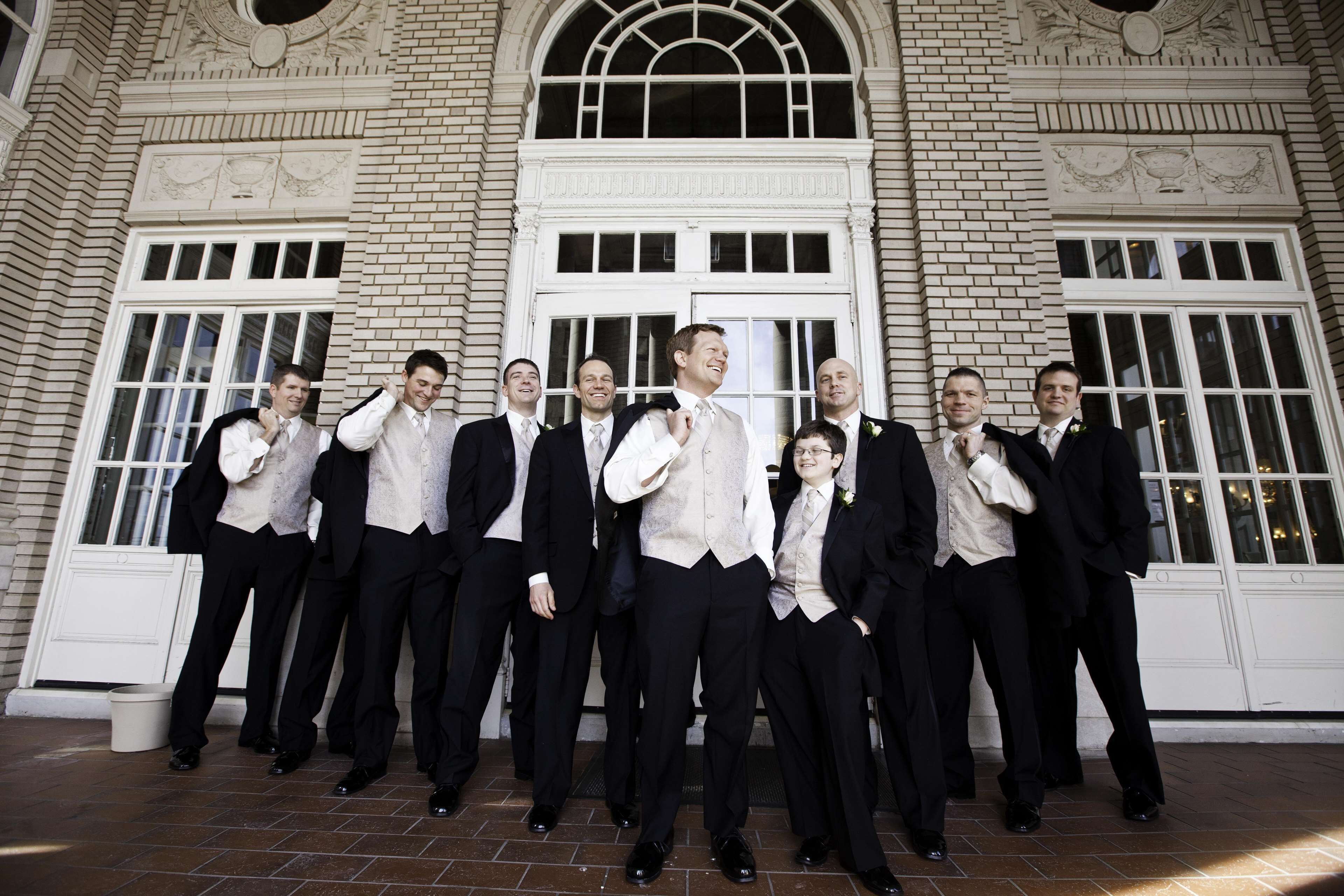 A group of men in formal attire standing in front of a building with large windows and decorative architectural elements.