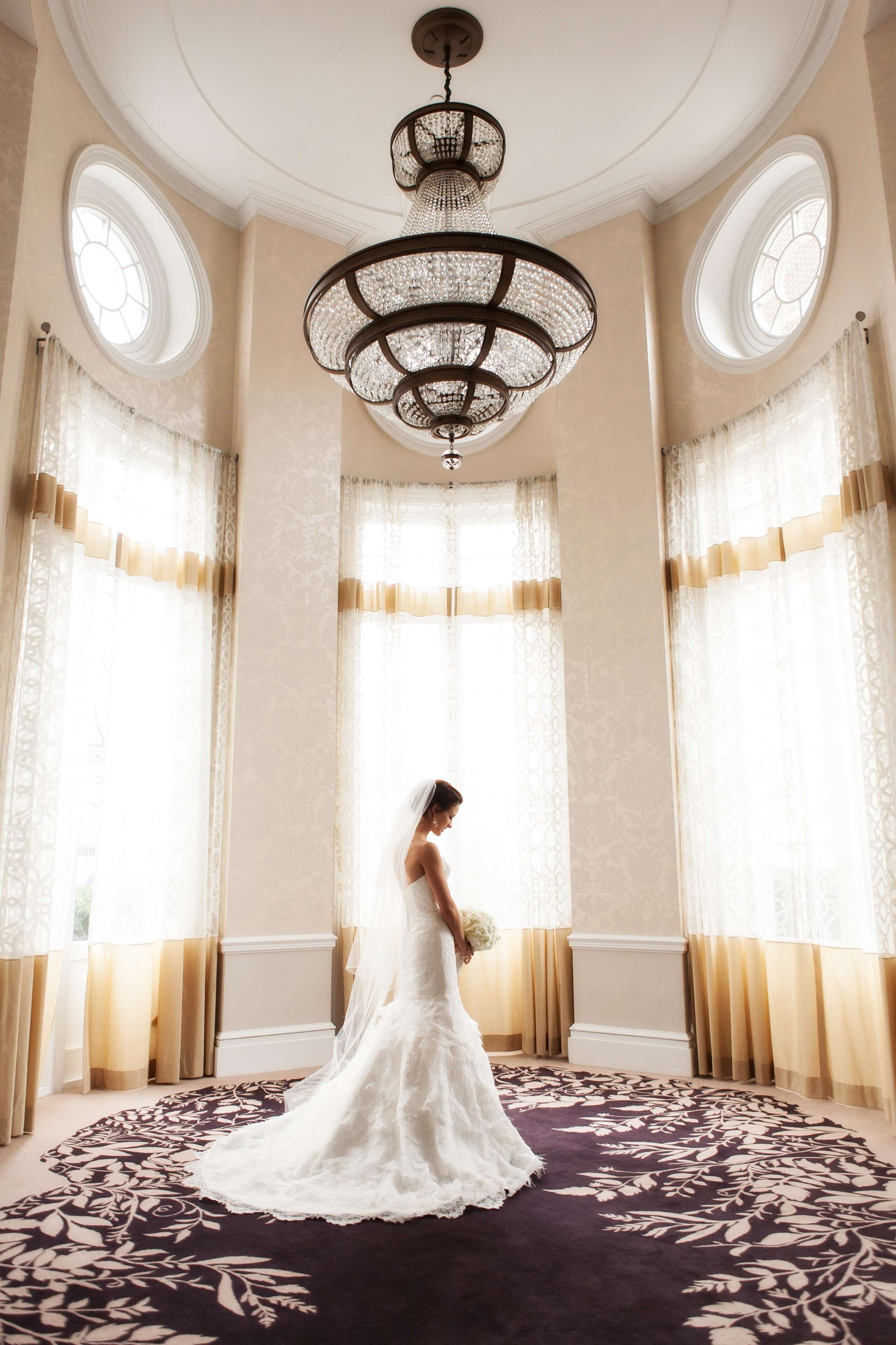 Une mariée, vêtue d’une robe de mariée blanche, se tient dans une pièce dotée de grandes fenêtres, d’un lustre et d’un tapis à motifs.