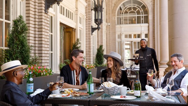 Un groupe de personnes est assis à une table en plein air, devant un bâtiment historique, et y prend son repas.