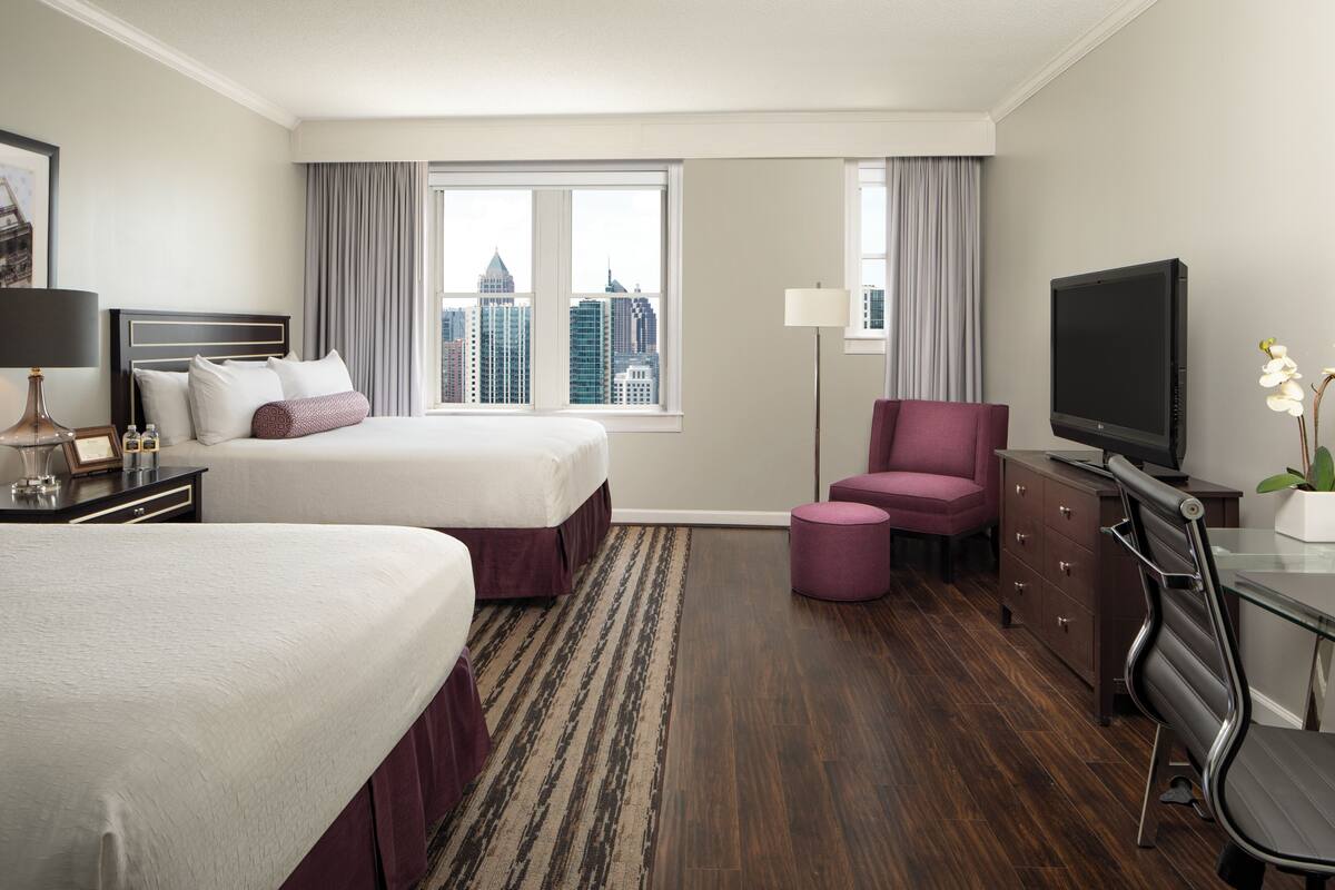 A hotel room with two beds, a desk with a TV, a chair, a dresser, and a view of the cityscape.