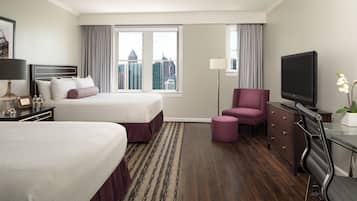 A hotel room with two beds, a desk with a TV, a chair, a dresser, and a view of the cityscape.