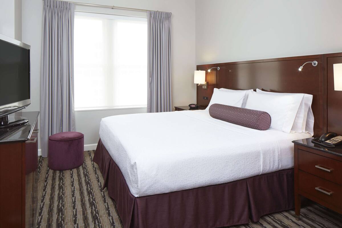 A hotel room with a large bed, a TV, a nightstand, and a window with curtains.