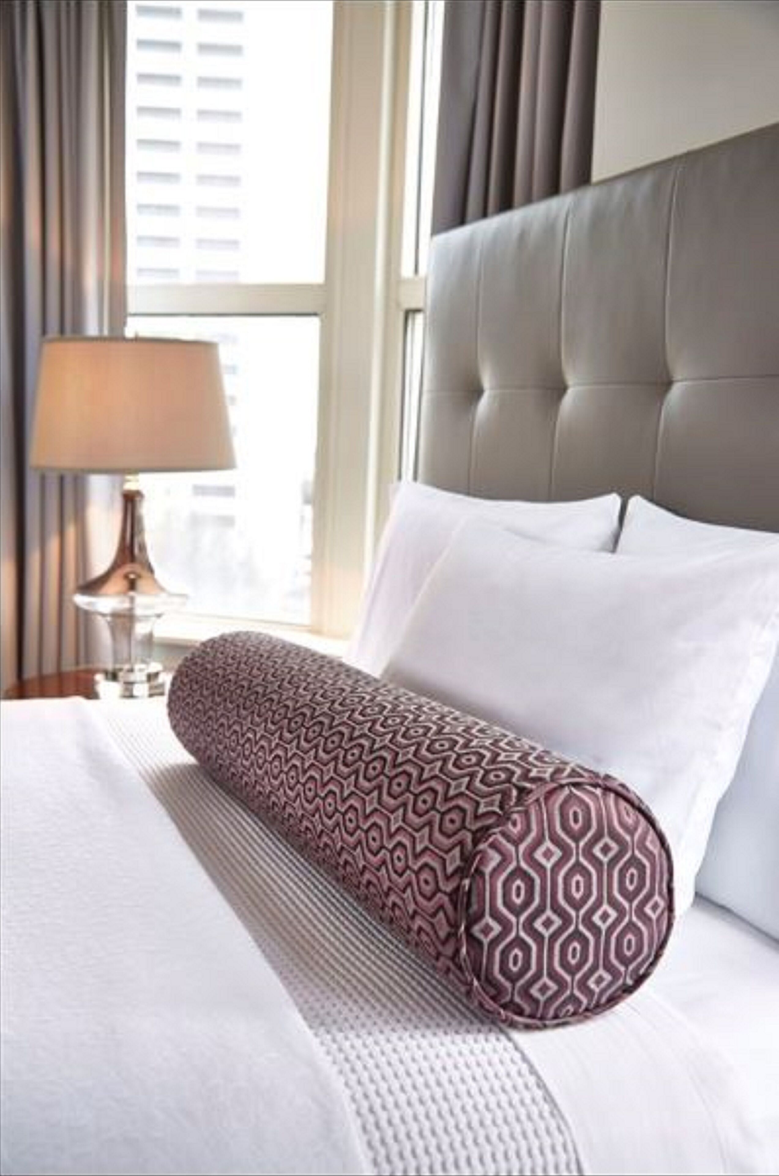 A hotel room with a tufted headboard, a patterned pillow, a lamp, and a window with curtains.