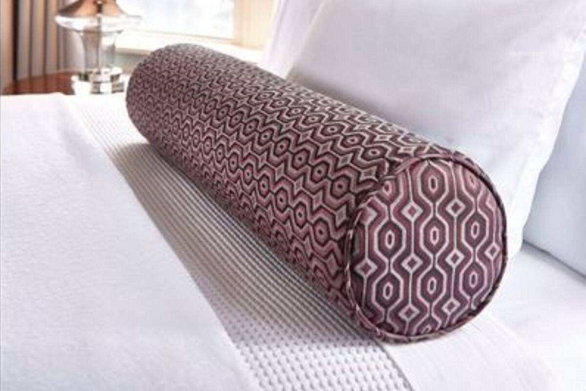 A hotel room with a tufted headboard, a patterned pillow, a lamp, and a window with curtains.