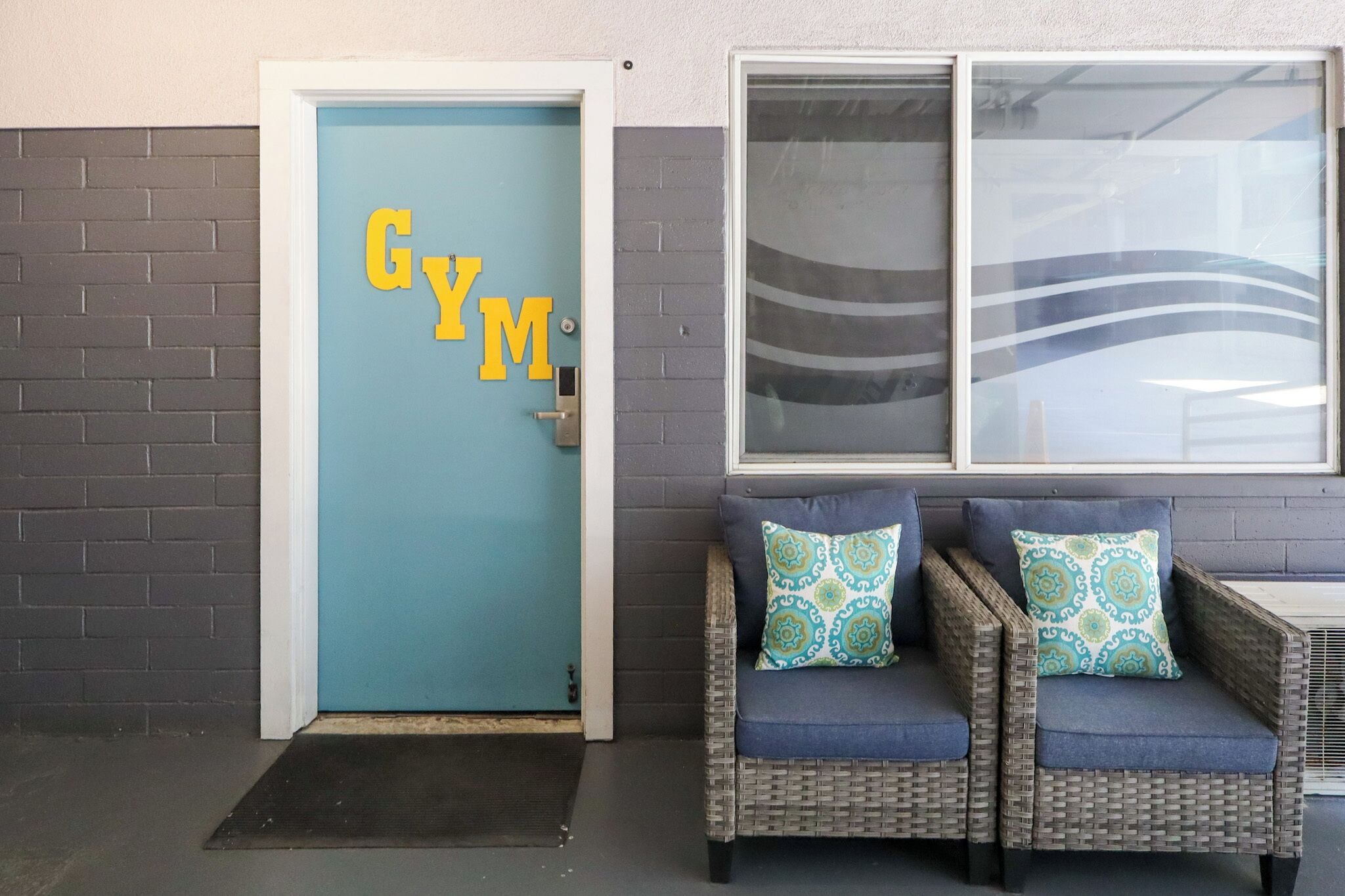 Una puerta con inscripción 'GYM', un sofá de mimbre con cojines estampados y una ventana con un diseño ondulado.