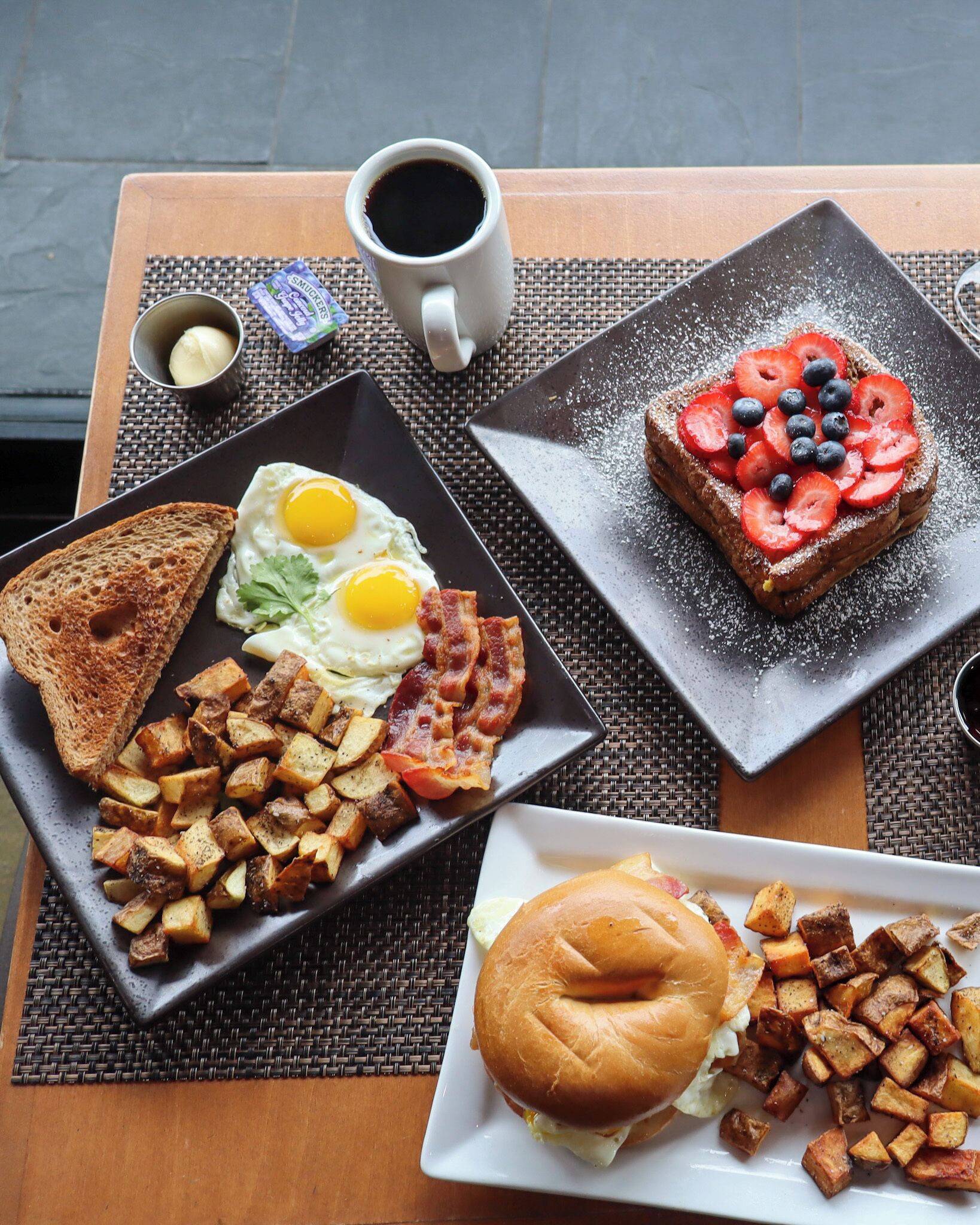 A breakfast table with toast, eggs, bacon, potatoes, a sandwich, and a cup of coffee.